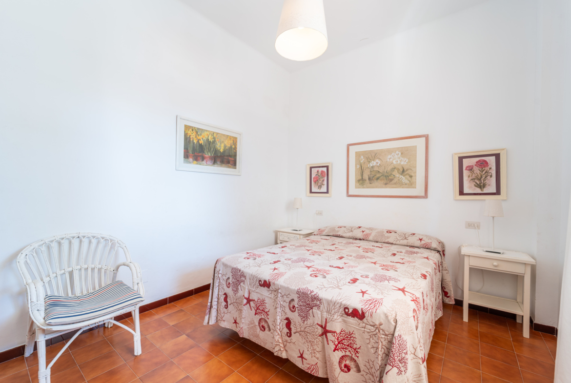Dormitorio con cama doble, butaca de mimbre blanca y cuadros enmarcados.