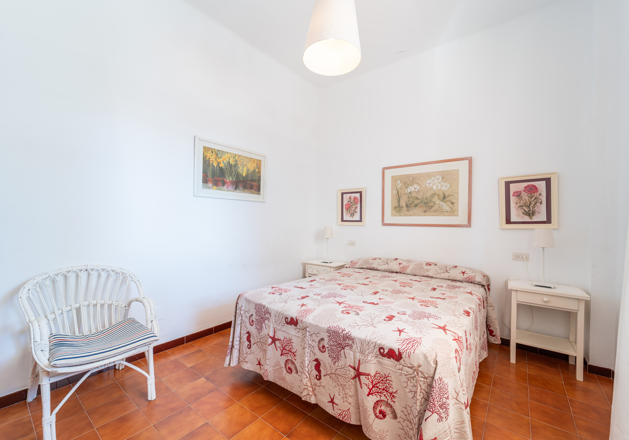 Dormitorio con cama doble, butaca de mimbre blanca y cuadros enmarcados.